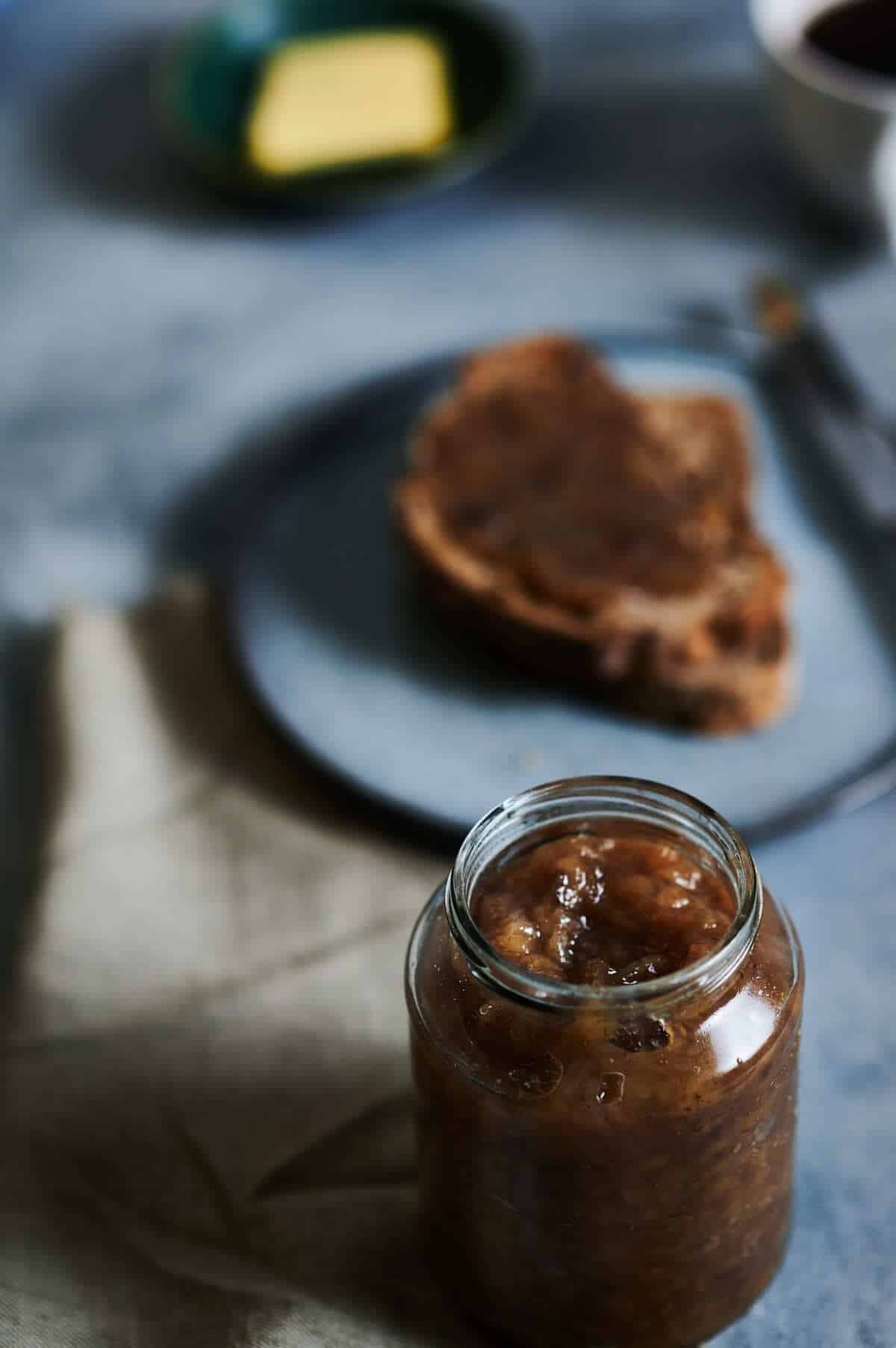 a glass jar filled with a dark brown jam with a piece of toast behind it