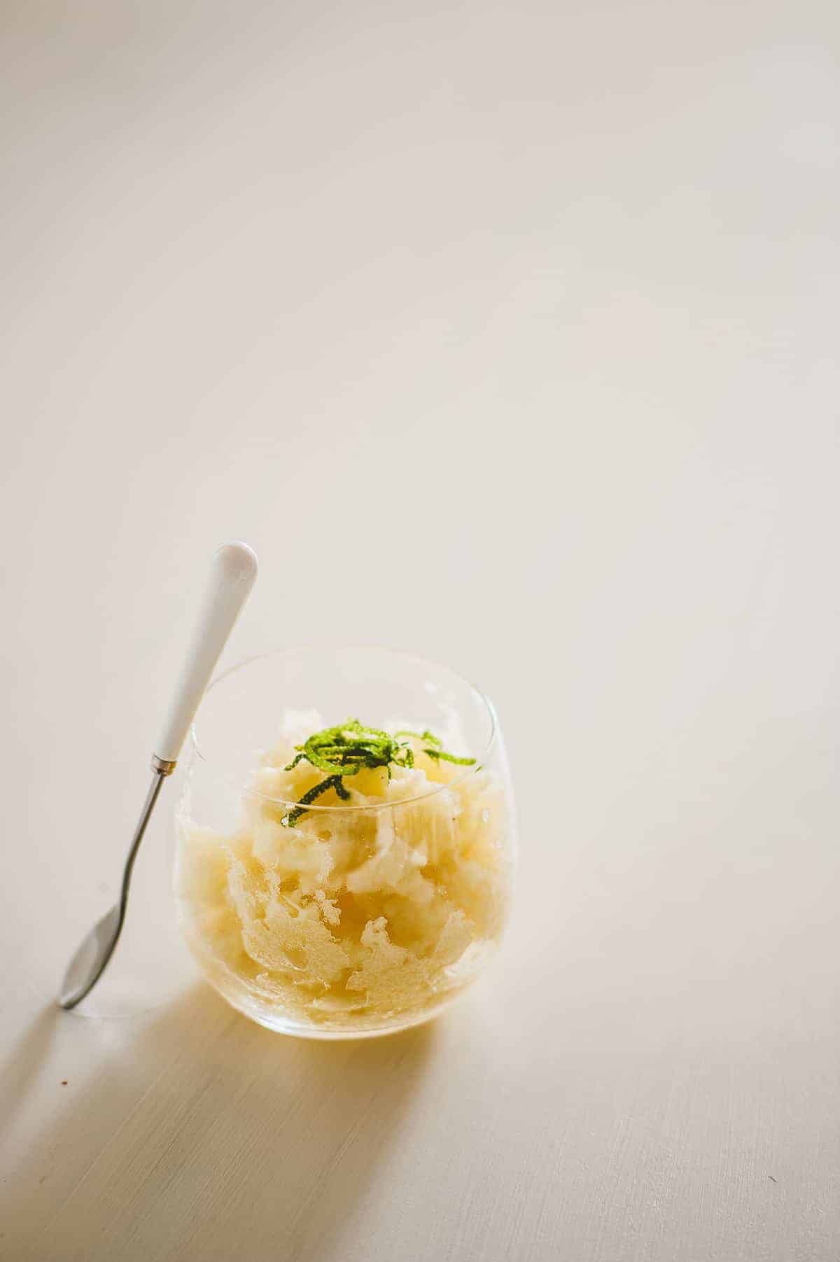 a glass on a white board filled with homemade pineapple granita with a spoon leaning against it