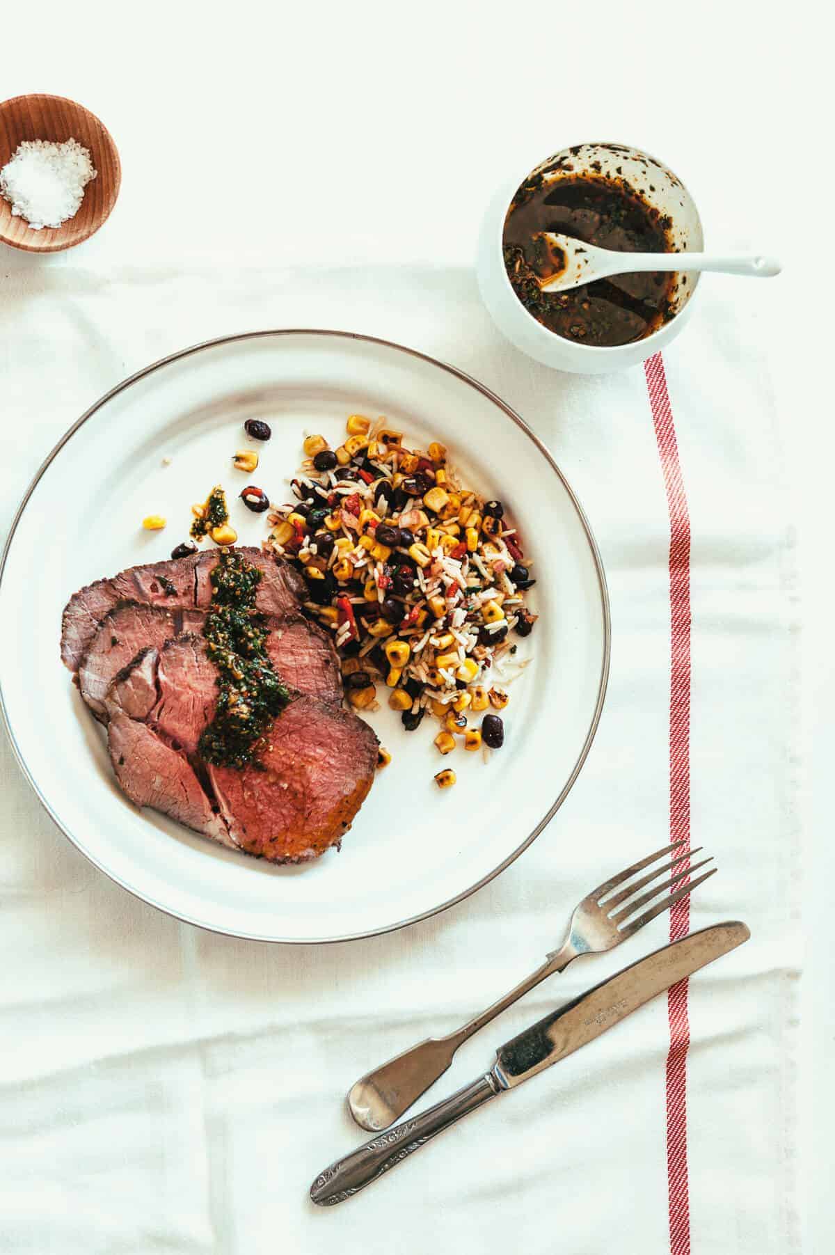 sliced roast meat served in a white plate with rice, beans and corn