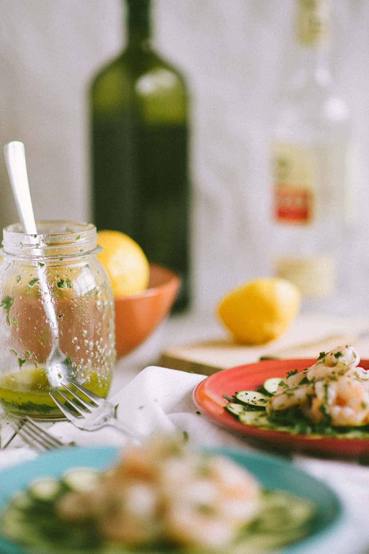 a jar filled with salad dressing on table with lemons and olive oil and two plates filled with food