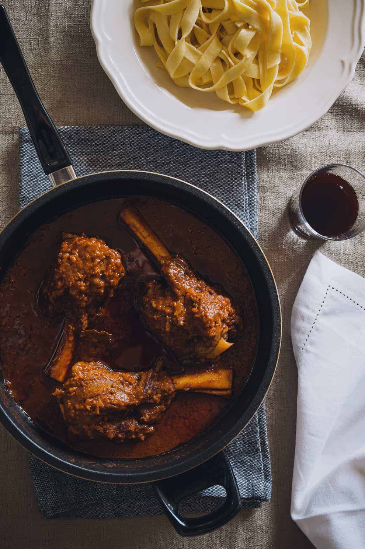 top down view of a pan containing three braised lamb shanks and a bowl of noodles