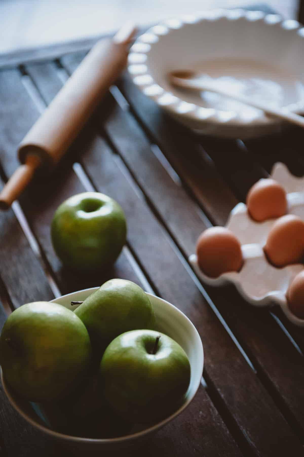 apples, eggs, a rolling pin and a dish too make apple pie on a wooden table