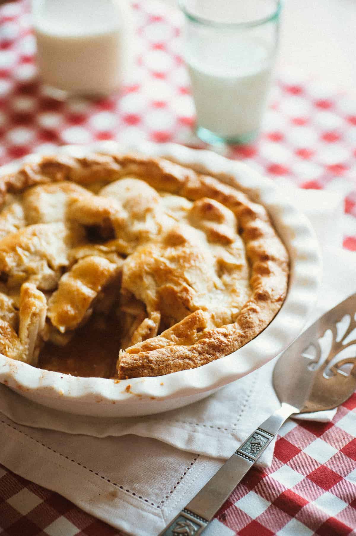 a baking dish with an apple pie on a table with a glass of milk