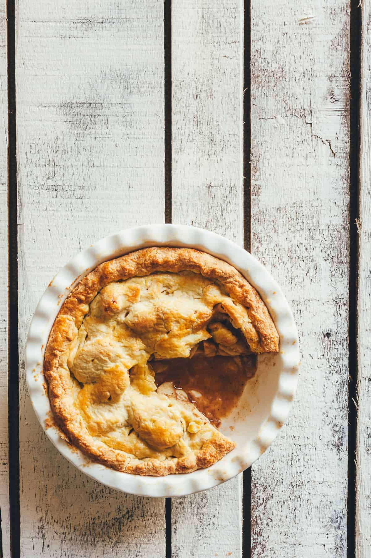 an apple pie in a baking dish with a slice cut from it on a white wooden table