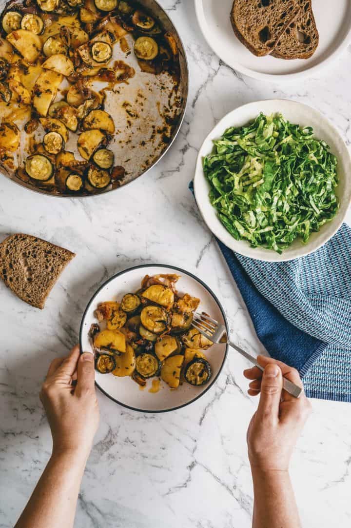 a top down view of a pair of hand eating from a plate with a baking dish of cooked food and a salad