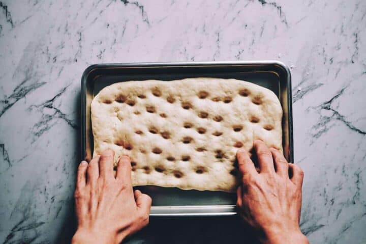 two hands making dimples in a shaped dough in a baking tray