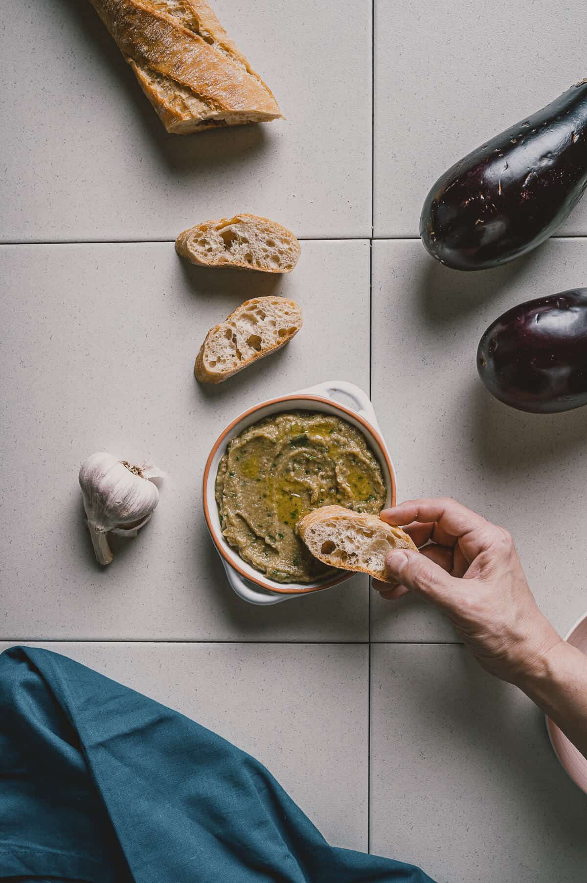 a hand scooping bread into a bowl with a kitchen towel nearby and 2 eggplants with garlic.