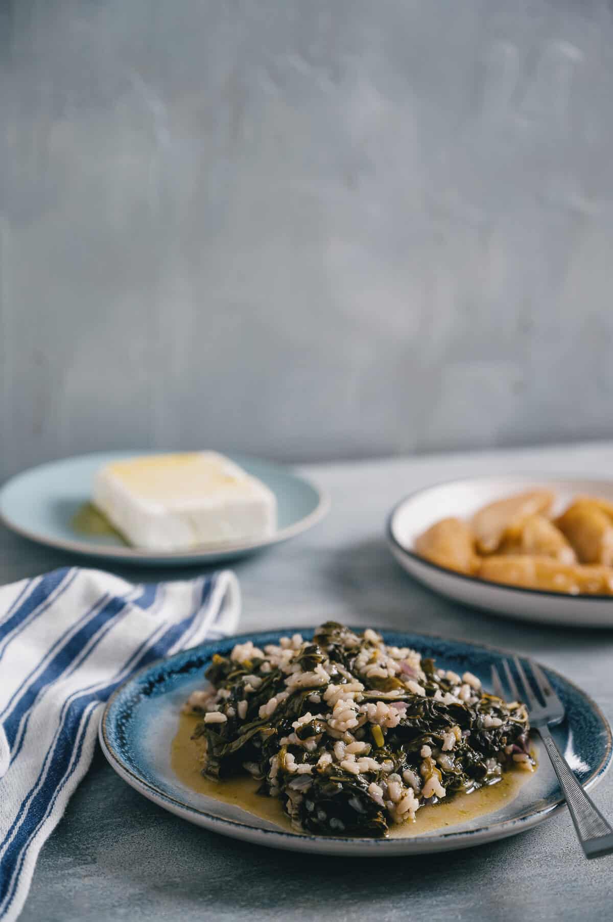 a plate of spinach cooked with rice on a grey table with a side of roast potatoes and feta cheese.