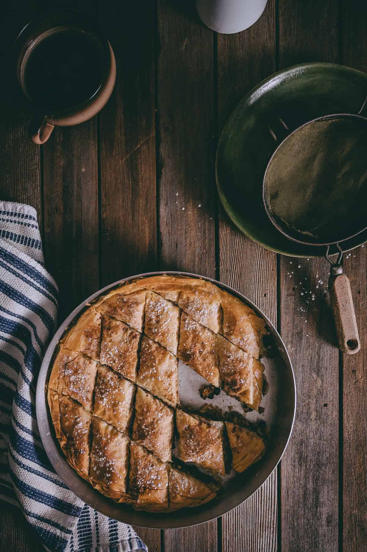 a filo pie on a wooden table with a kitchen towel and a cup of coffee.