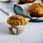 pumpkin banana muffin in a white parchment wrapper on a table in front of a blue plate of other muffins.