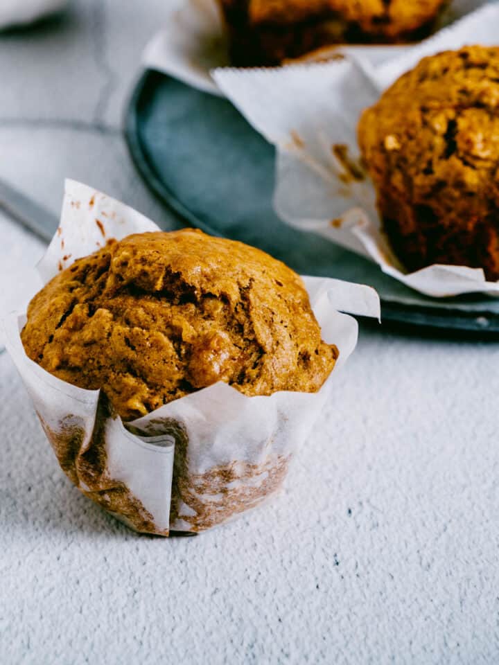 pumpkin banana muffin in a white parchment wrapper on a table in front of a blue plate of other muffins.