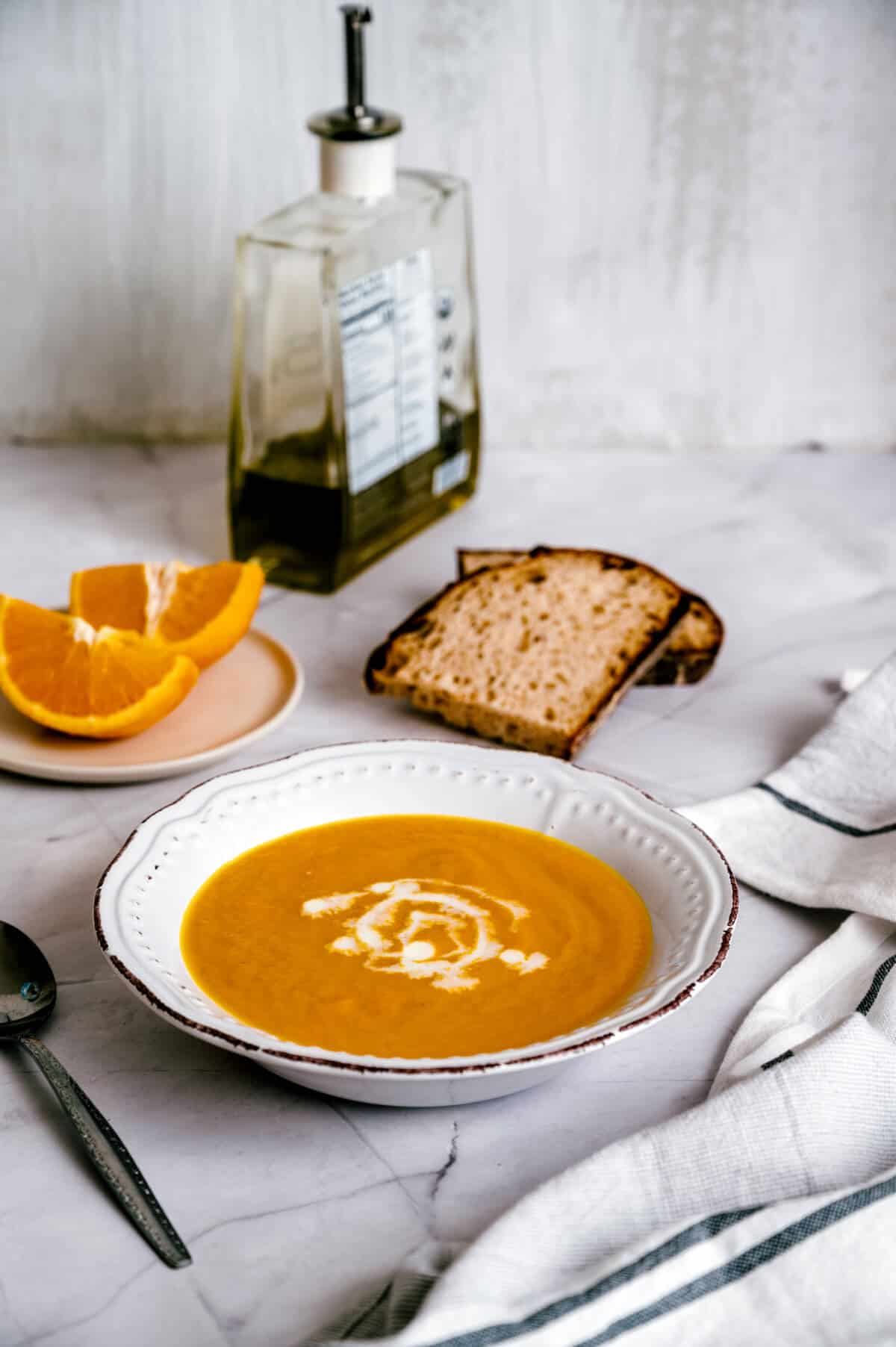 45 degree angle shot of a white bowl of carrot orange soup with a plate of orange segments and two slices of toast in the background.