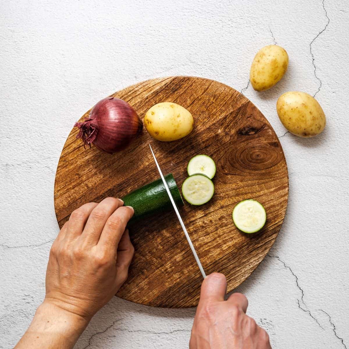 chopping zucchini, potatoes and onions on a round cutting board.
