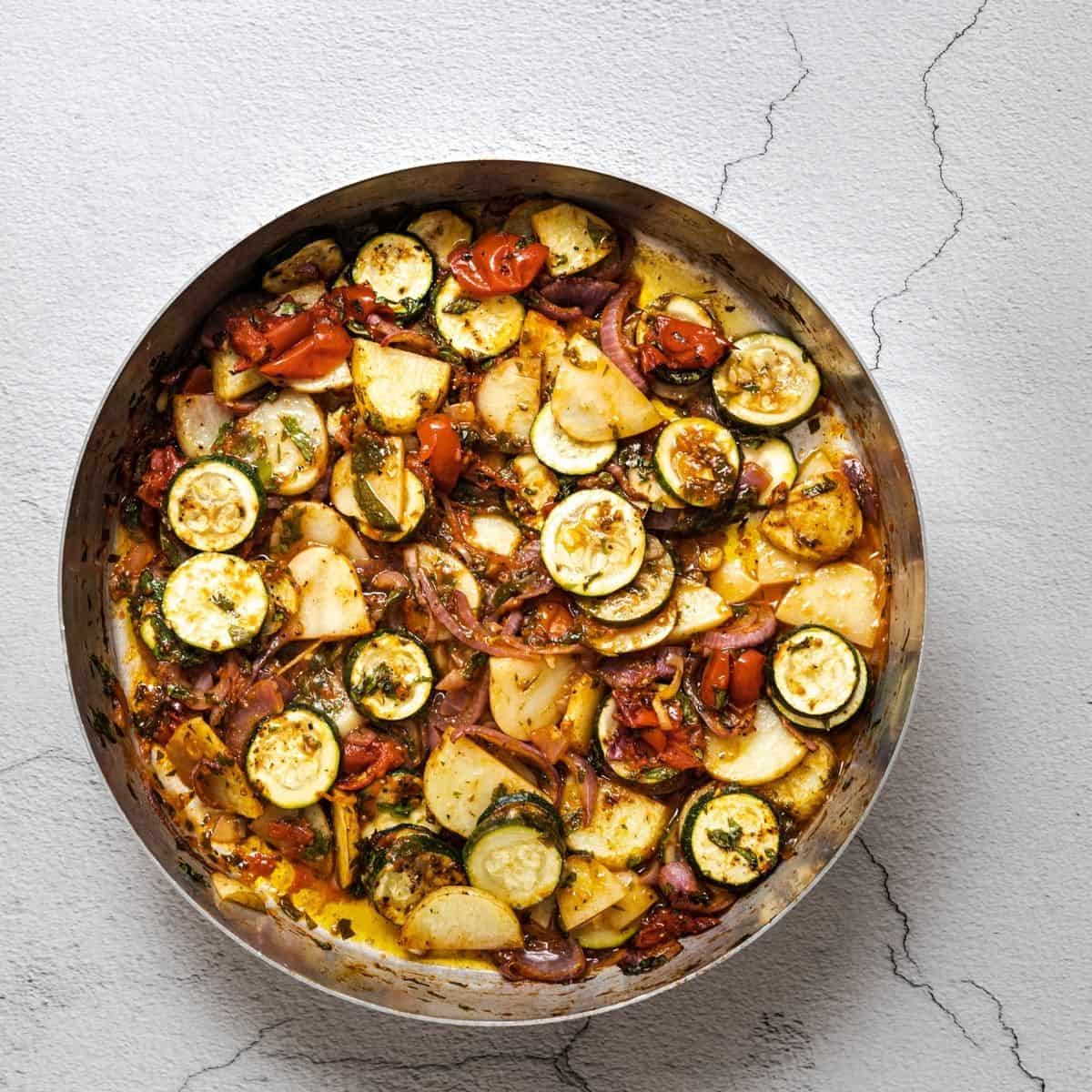 a round baking dish filled with Greek roasted vegetables on a white concrete surface.