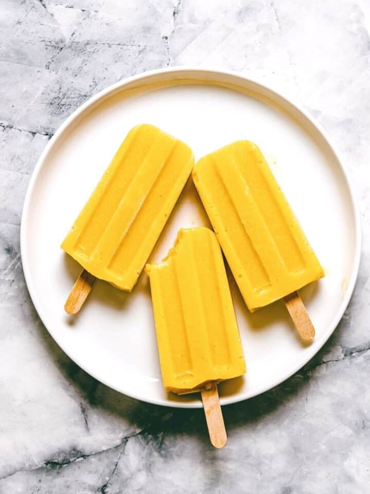 3 mango popsicles on a white plate on a marble surface.