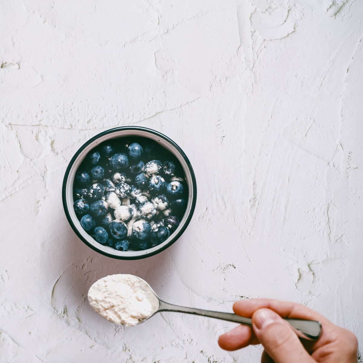 a hand spooning flour over blueberries in a bowl.