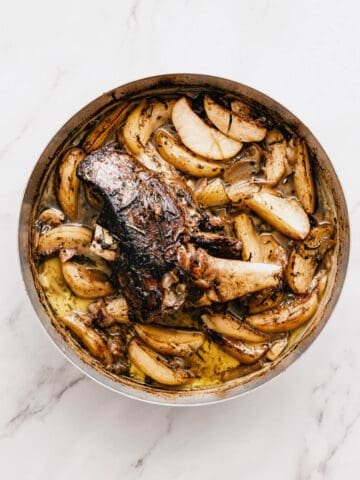a roast leg of lamb with potatoes in a round roasting pan on a white table.