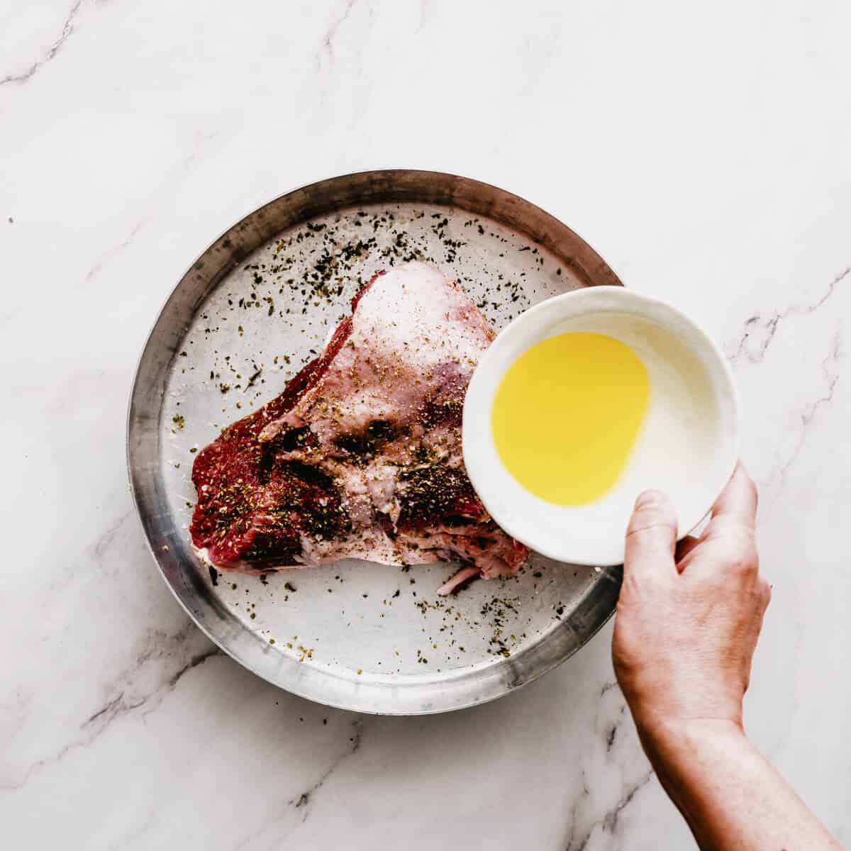 pouring olive oil over a leg of lamb in a round roasting pan.