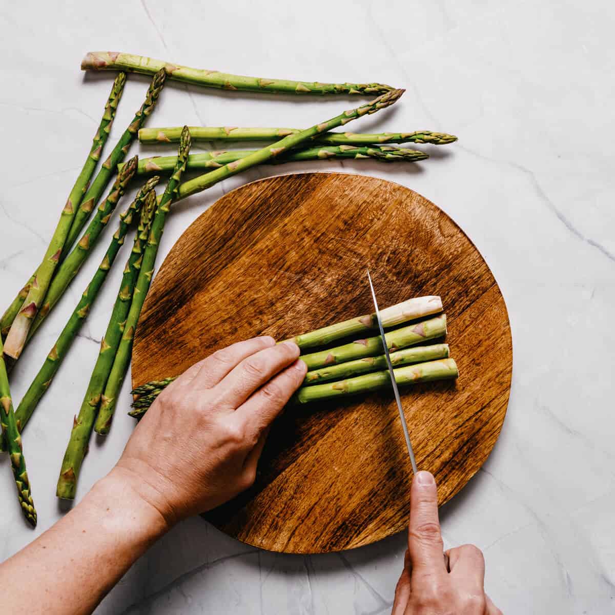 asparagus spears being cut with a knife on a round wooden chopping board.