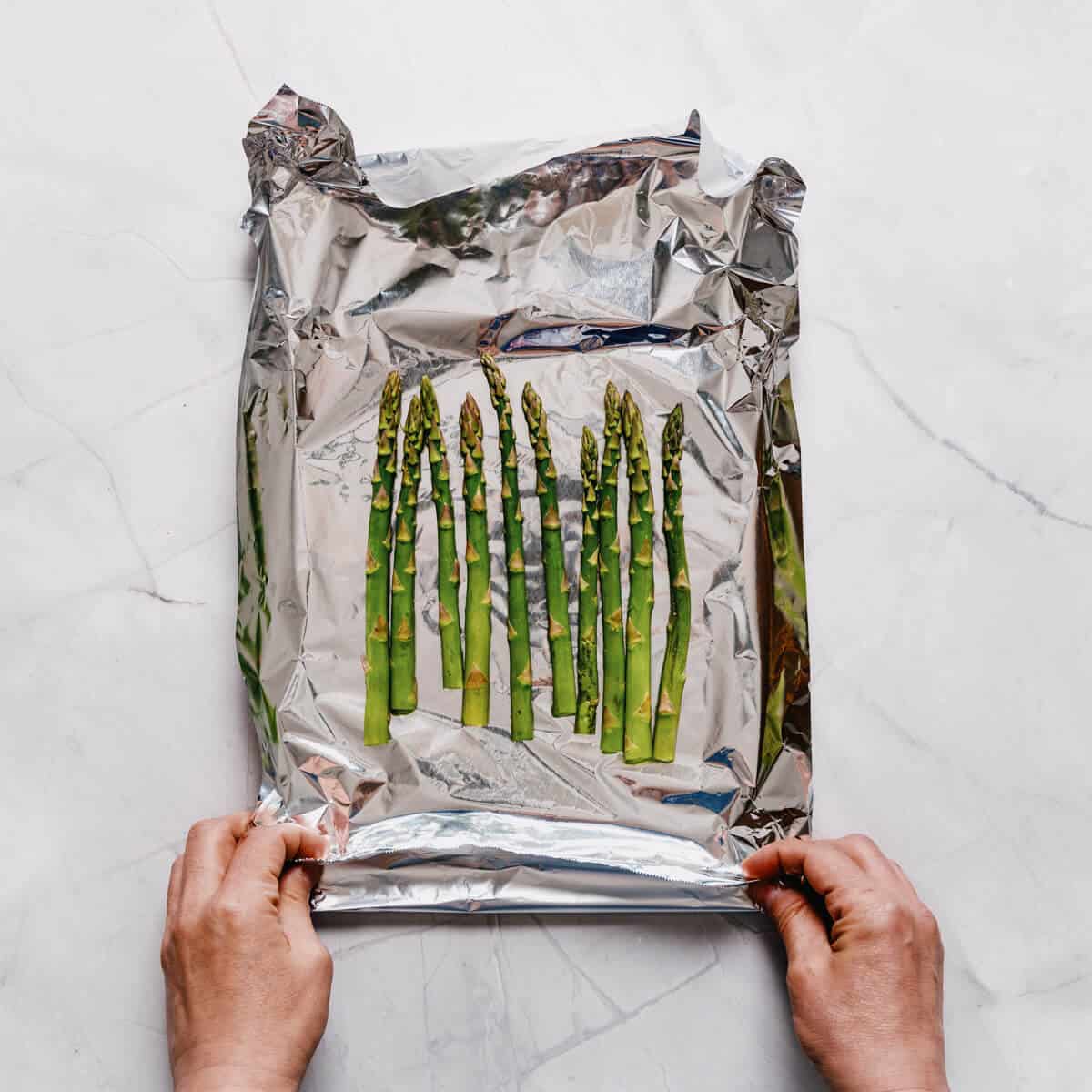a pair of hands sealing an aluminum foil packet filled with asparagus.