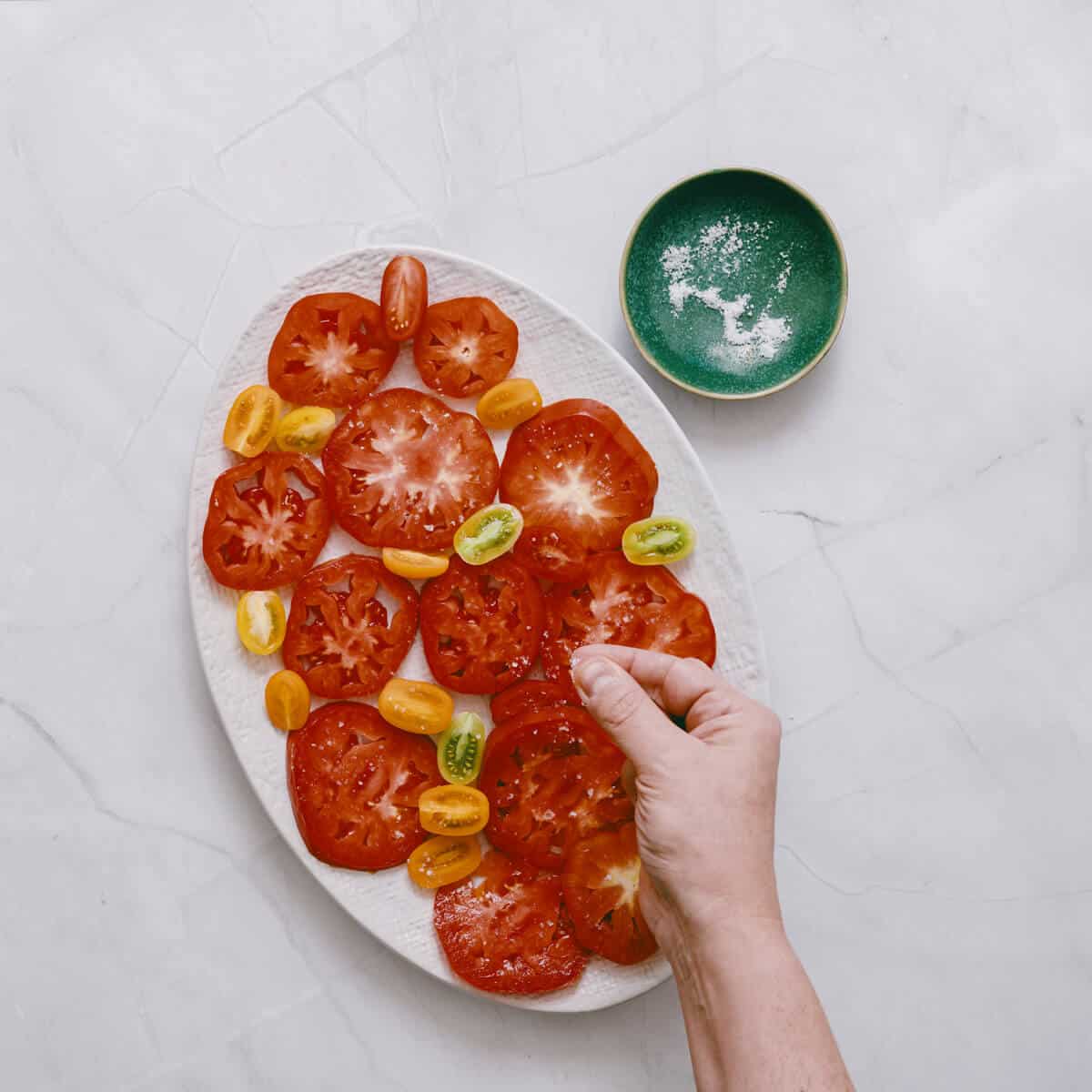 sprinkling salt on sliced heirloom tomatoes on a white platter.