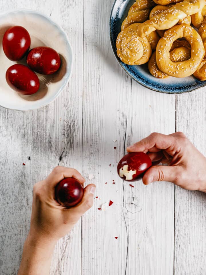 two hands cracking Greek Easter eggs with cookies on a white wooden table.
