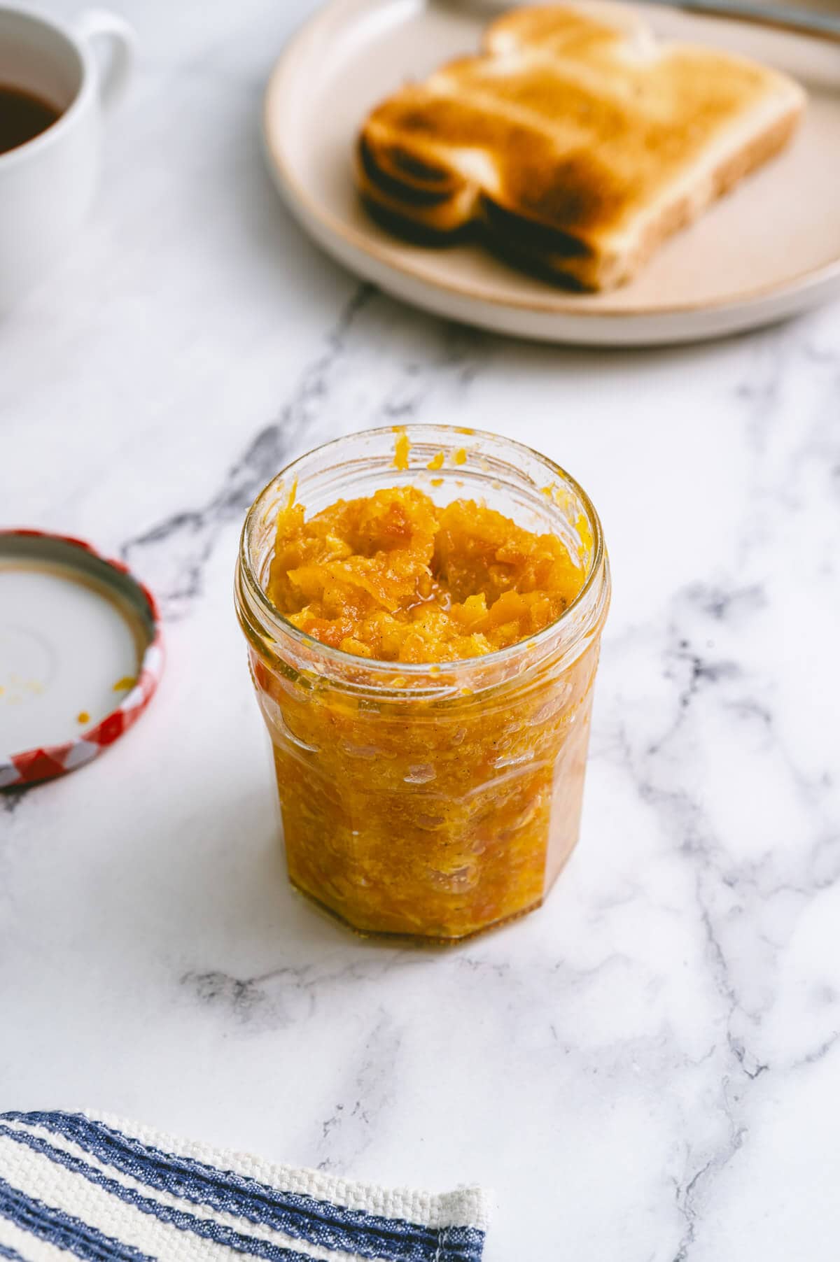 an opened jar of mandarin jam on a table served with a slice of toasted bread.