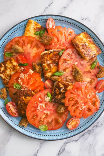 Blue plate with tomato and halloumi salad, topped with croutons and fresh basil.