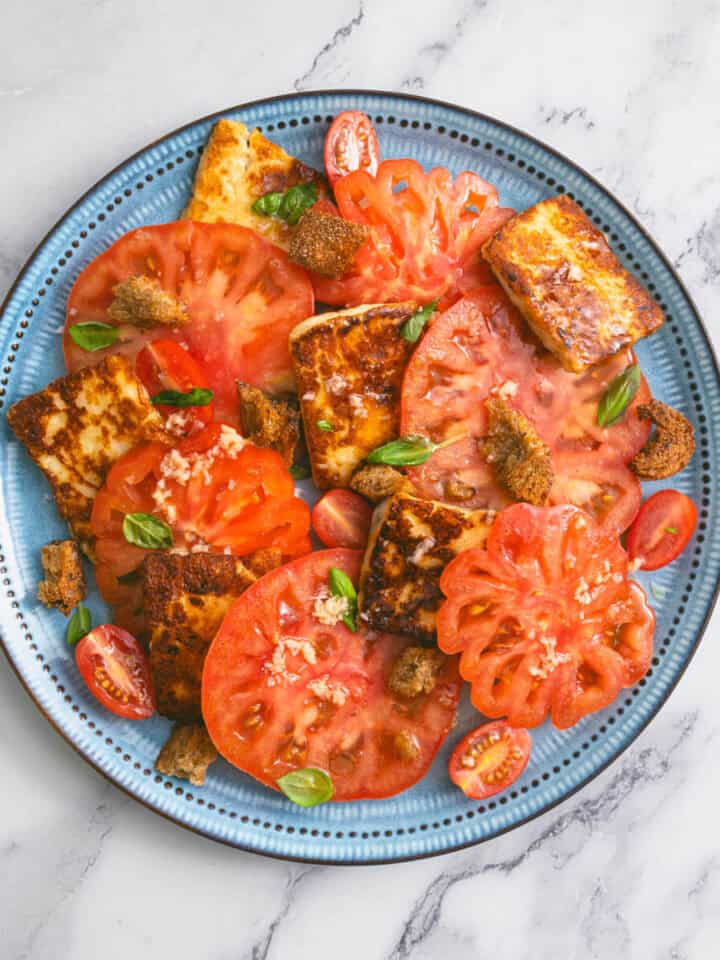 Blue plate with tomato and halloumi salad, topped with croutons and fresh basil.