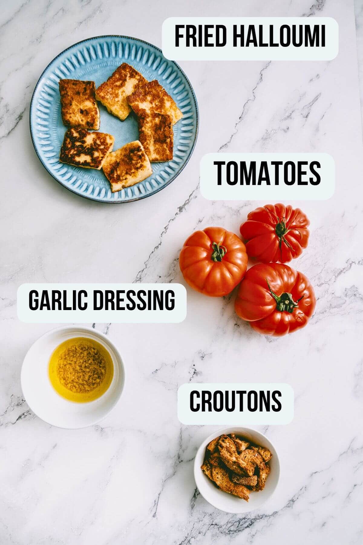 Flat lay of ingredients for tomato and halloumi salad including fried halloumi, tomatoes, garlic dressing, and croutons.