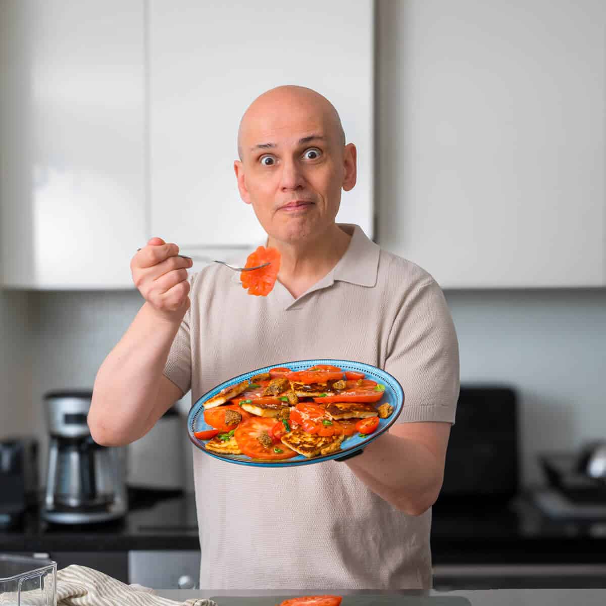 Man holding a plate of tomato and halloumi salad, smiling in the kitchen.