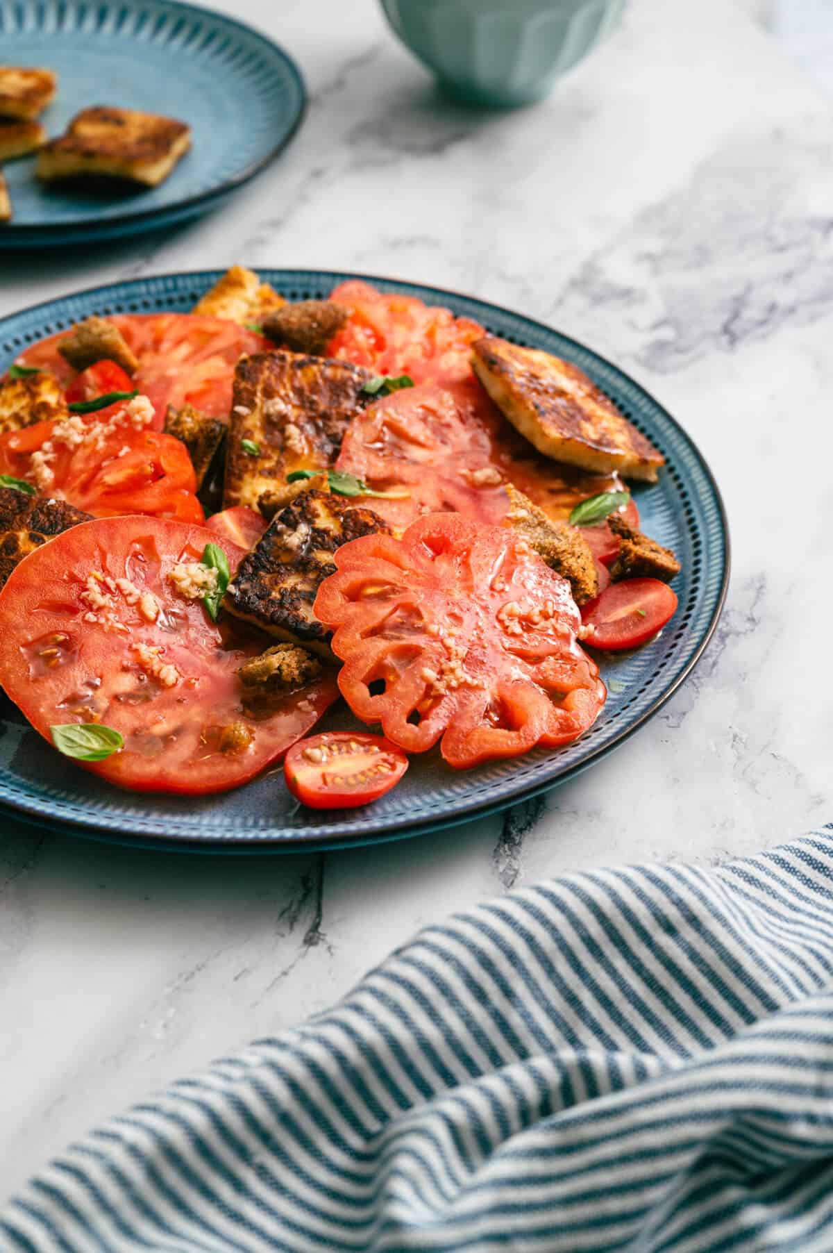 Side view of tomato and halloumi salad with striped cloth napkin on marble table.