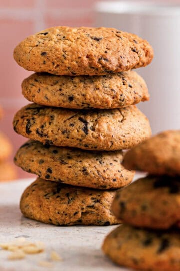 Stack of chocolate almond cookies showing their golden texture and chocolate flecks, with more cookies blurred in the background on a soft pink surface.