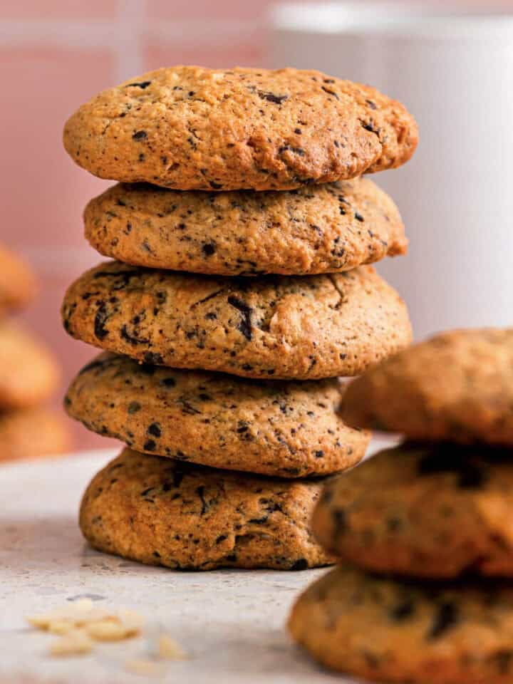 Stack of chocolate almond cookies showing their golden texture and chocolate flecks, with more cookies blurred in the background on a soft pink surface.