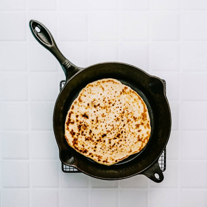 Greek pita bread cooking in a hot cast iron pan with golden spots forming.