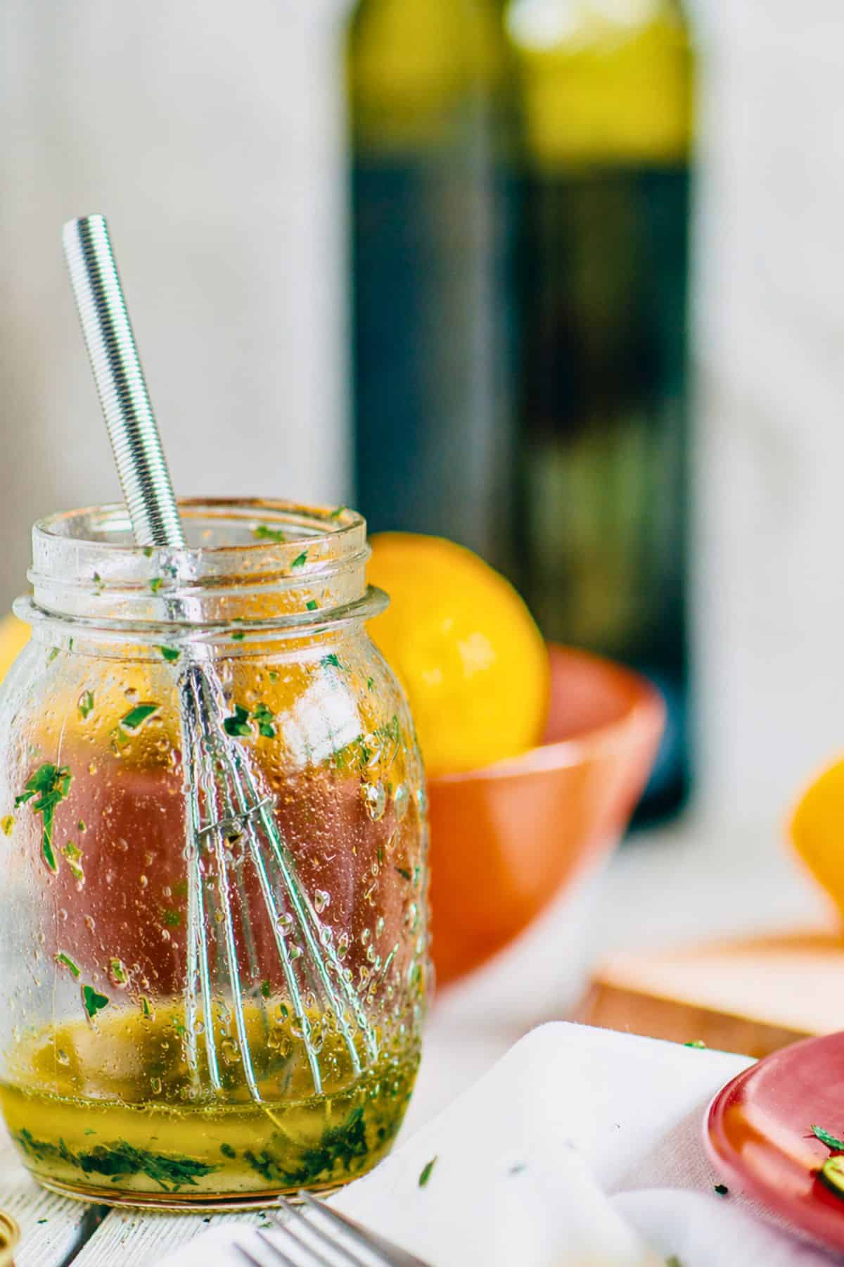 Close up of Greek salad dressing with ouzo in a glass jar with herbs, lemon and olive oil in the background.