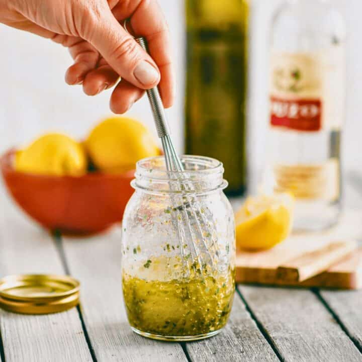 Greek salad dressing with ouzo being mixed in a glass jar with a whisk and fresh herbs.