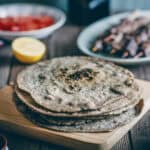 a stack of buckwheat pita breads on a wooden board with souvlaki sticks and tomatoes in the background.