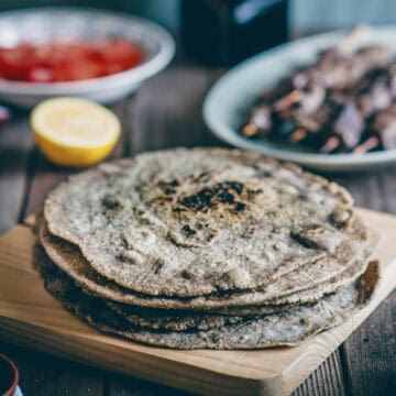 a stack of buckwheat pita breads on a wooden board with souvlaki sticks and tomatoes in the background.