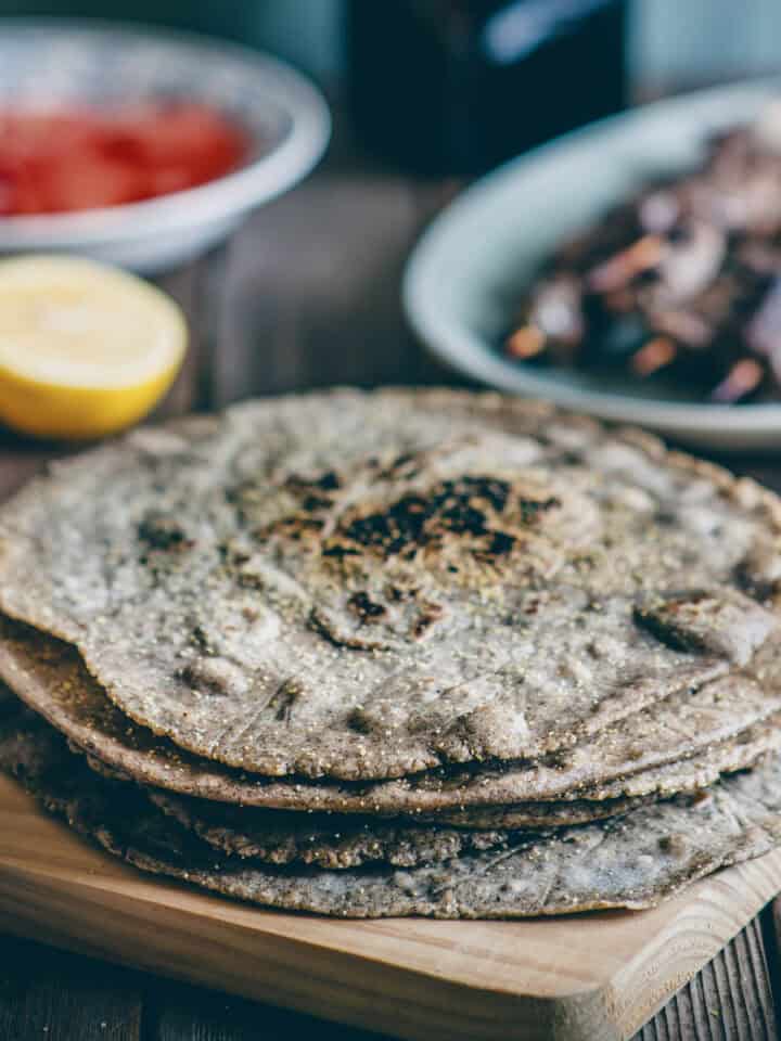 a stack of buckwheat pita breads on a wooden board with souvlaki sticks and tomatoes in the background.