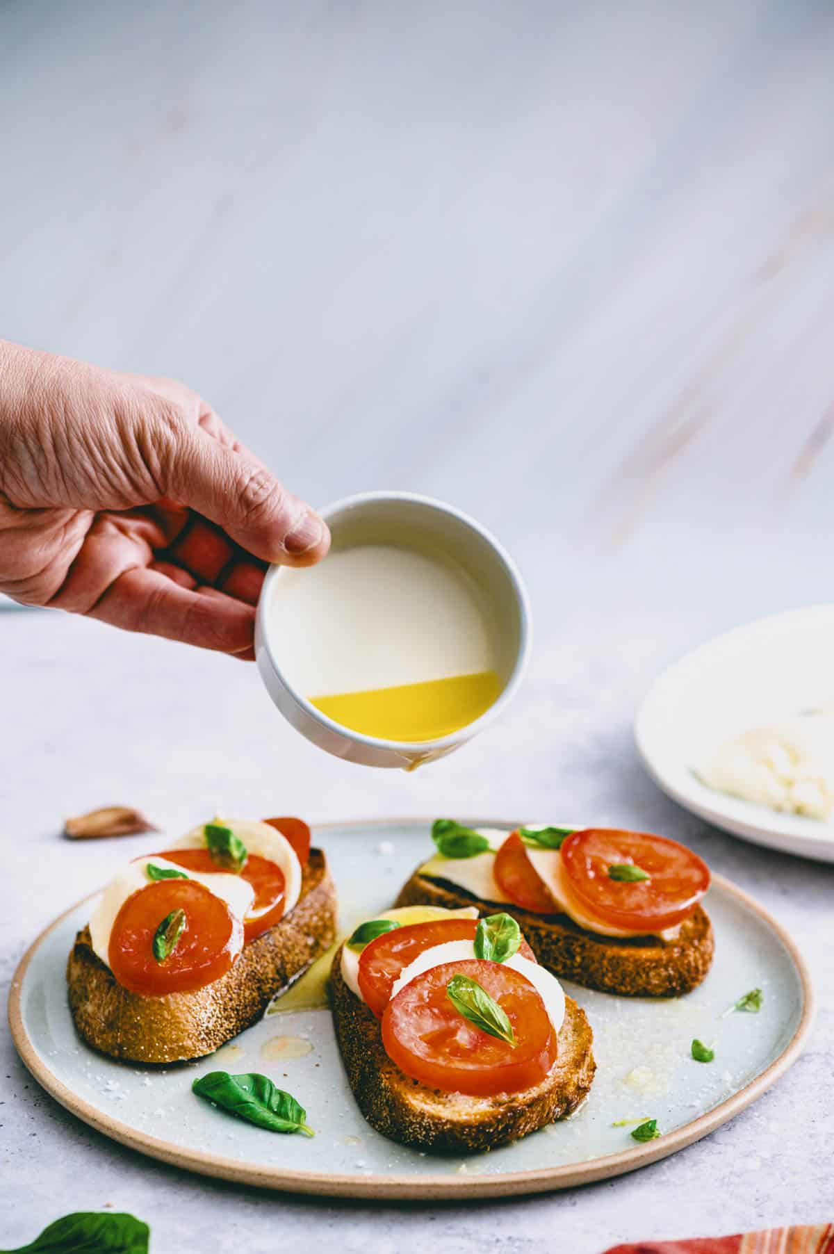 A hand pours olive oil from a small white bowl onto slices of bread topped with mozzarella, tomato, and fresh basil, arranged on a round plate.