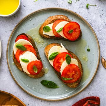 Three slices of toasted bread topped with fresh mozzarella, tomato slices, and basil leaves are arranged on a grey plate, drizzled with olive oil, and garnished with extra basil.