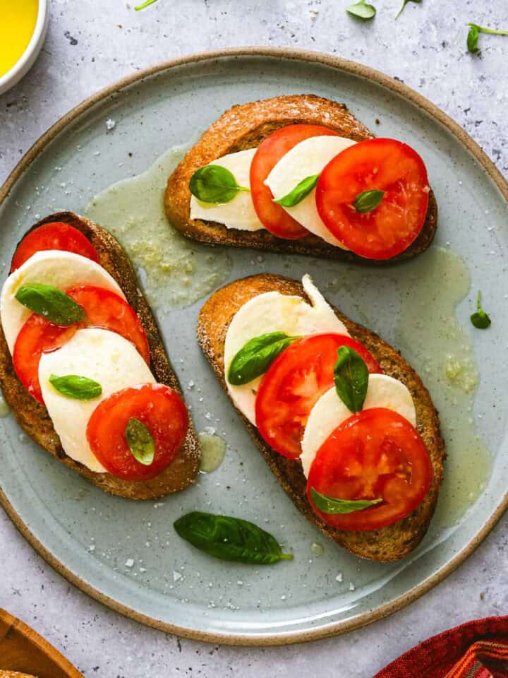 Three slices of toasted bread topped with fresh mozzarella, tomato slices, and basil leaves are arranged on a grey plate, drizzled with olive oil, and garnished with extra basil.