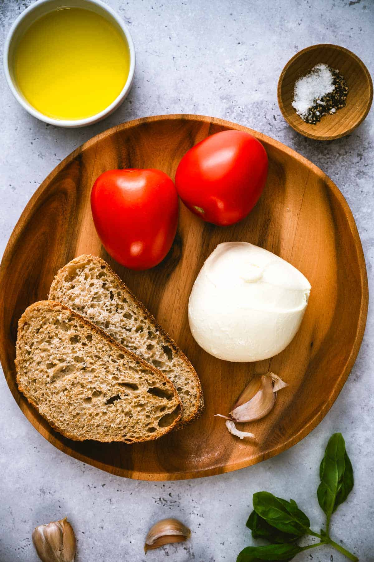 A wooden plate with two slices of bread, two Roma tomatoes, a ball of mozzarella, and garlic cloves. Surrounding the plate are a bowl of olive oil, salt and pepper, and fresh basil leaves on a light surface.