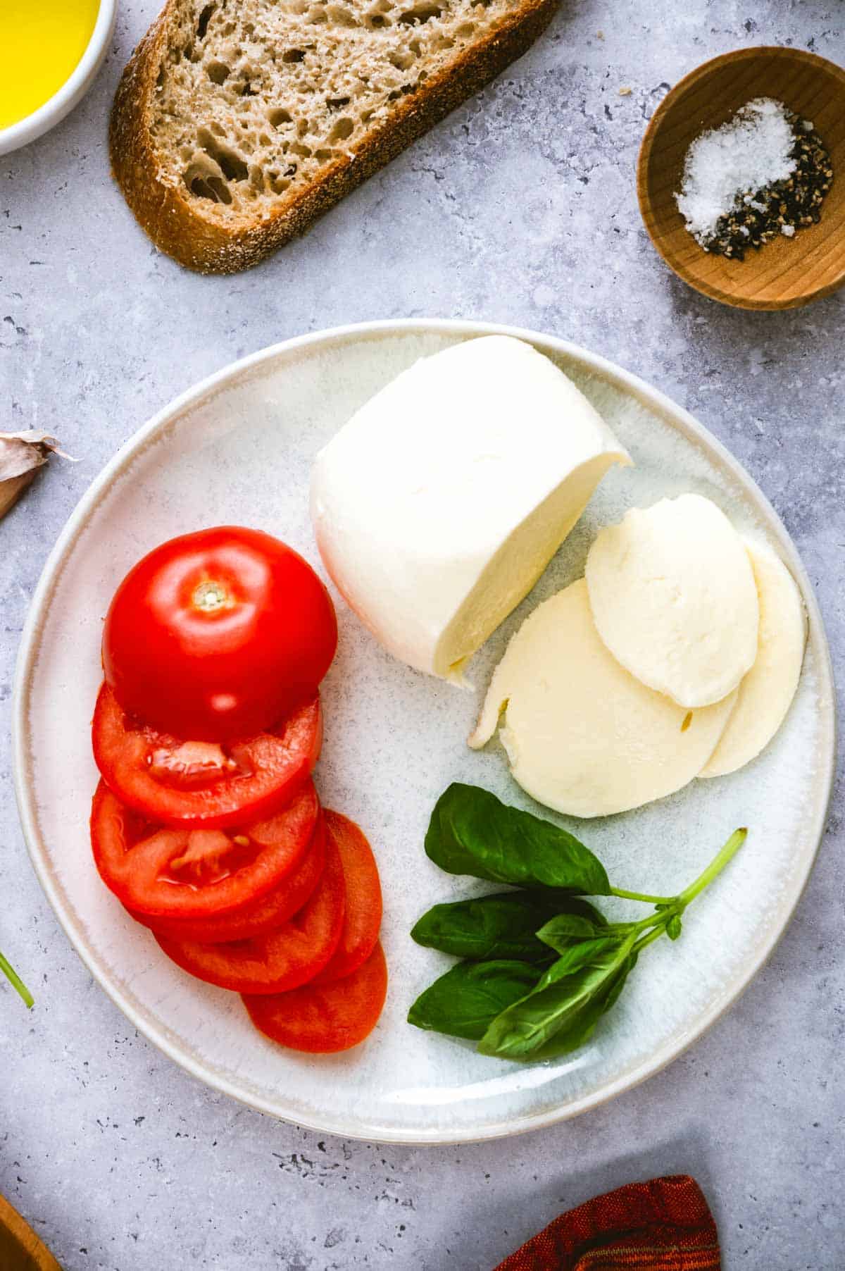A plate with sliced tomato, fresh mozzarella cheese (whole and sliced), and basil leaves. Nearby are a slice of bread, a small bowl of salt and pepper, and a small dish of olive oil on a gray surface.