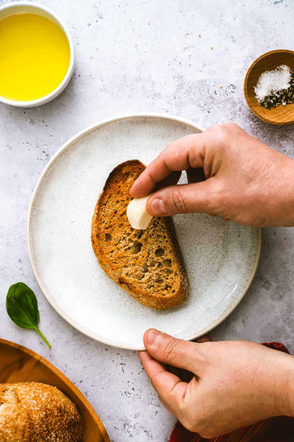 A person rubs a peeled garlic clove onto a slice of toasted bread on a white plate. Nearby are a bowl of olive oil, a wooden bowl of salt and pepper, and a fresh basil leaf.