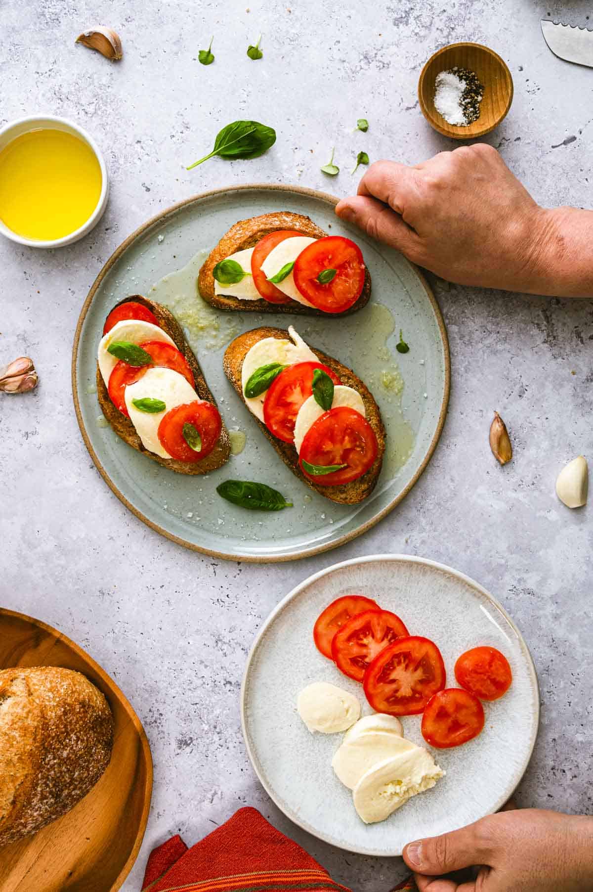 Three slices of bread topped with tomato, mozzarella, and basil are arranged on a plate. Nearby, a smaller plate has sliced tomato and mozzarella. Hands are holding the main plate, with olive oil and salt visible on the table.