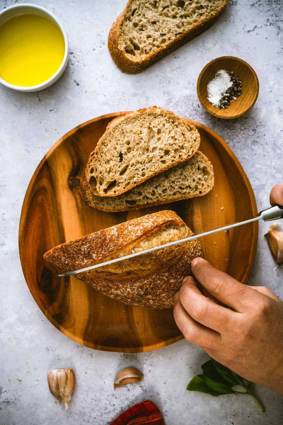 A hand slices a loaf of rustic bread on a wooden plate. Two slices are already cut. Nearby are a bowl of olive oil, a small dish of salt and pepper, garlic cloves, and some fresh herbs.