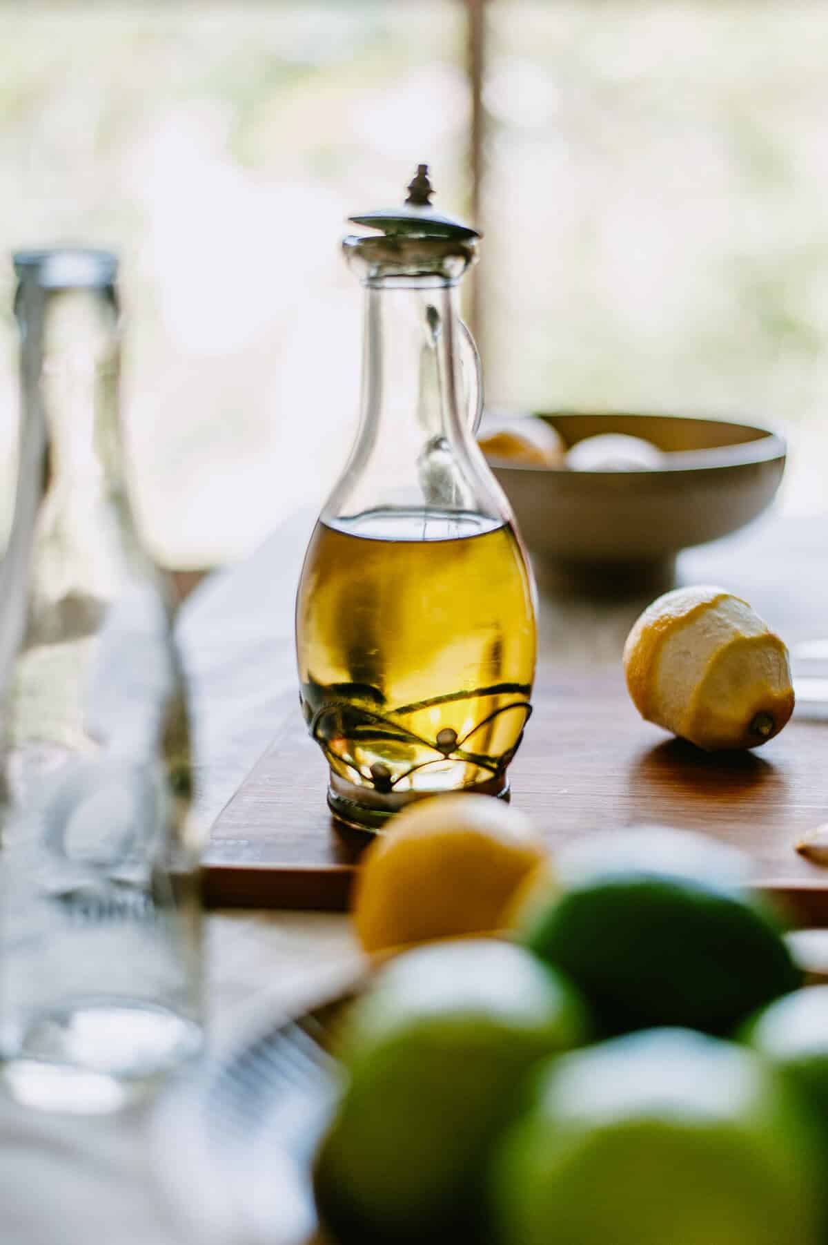 Glass bottle of lemon infused olive oil surrounded by lemons and limes on a wooden board.