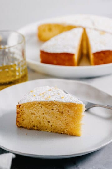 A slice of lemon ricotta cake made with olive oil on a white plate, with the whole cake and a glass of olive oil in the background.